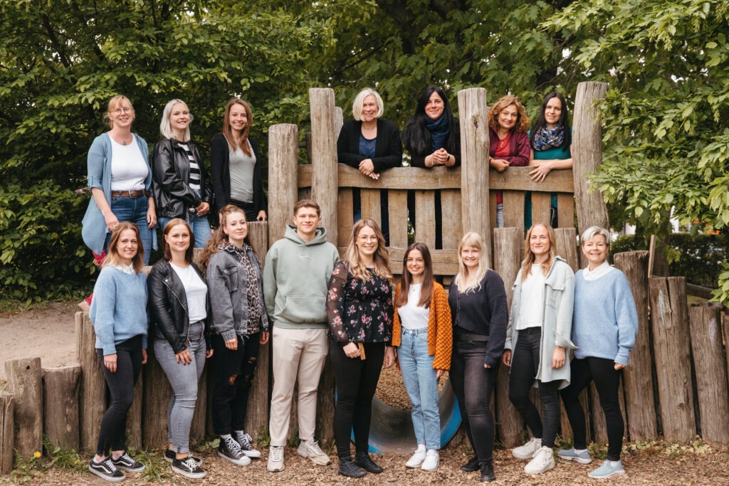 Team der Kita Sausewind in der Industriestraße 6 in Dresden, aufgenommen vor einem Holzspielplatz; eine Gruppe pädagogischer Fachkräfte in freundlicher und offener Atmosphäre im Grünen.