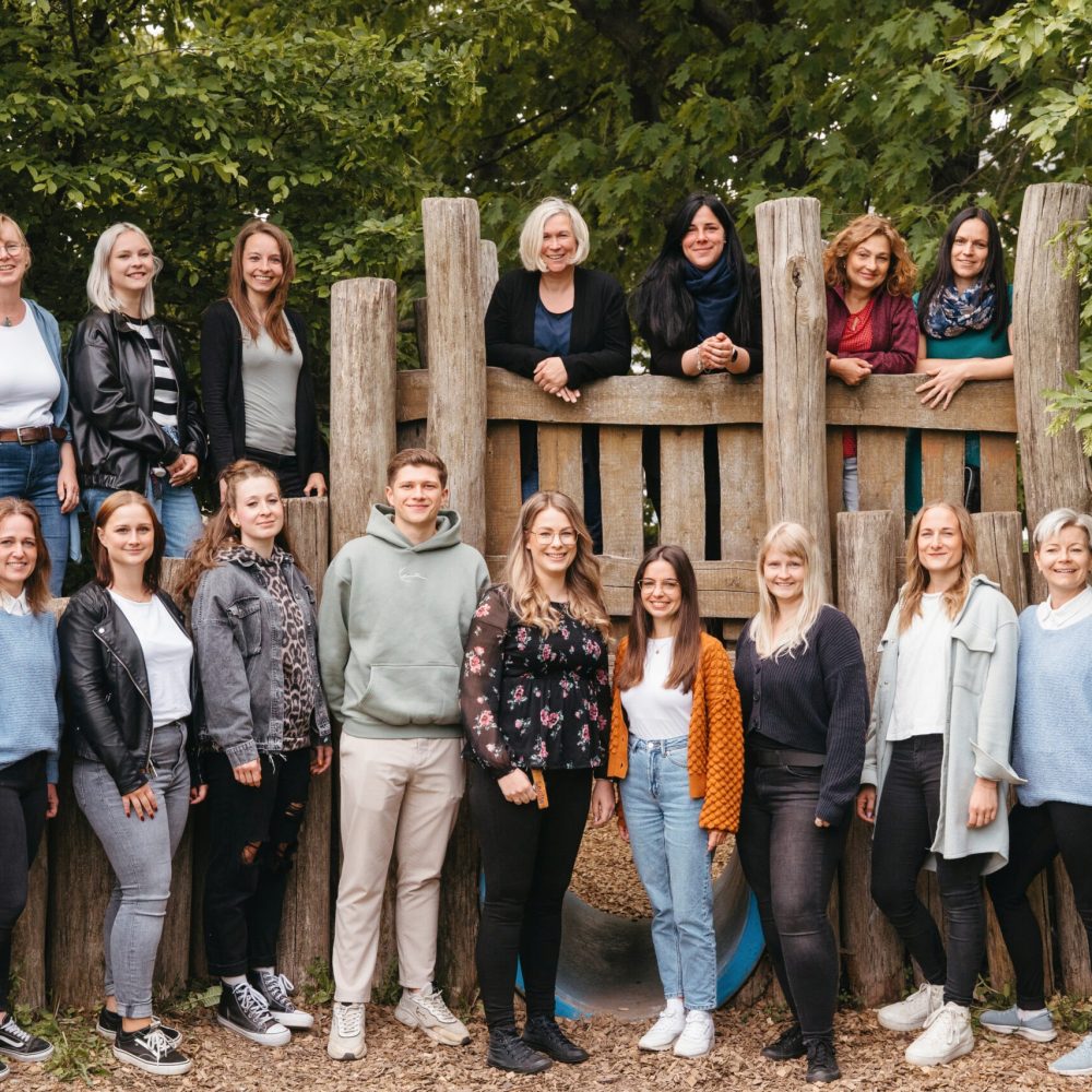 Team der Kita Sausewind in der Industriestraße 6 in Dresden, aufgenommen vor einem Holzspielplatz; eine Gruppe pädagogischer Fachkräfte in freundlicher und offener Atmosphäre im Grünen.