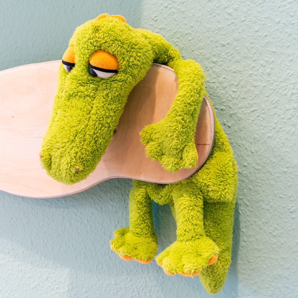Grünes Plüschkrokodil als Deko in einem Kita-Raum, entspannt über ein helles Holzbrett gehängt – freundliches, kindgerechtes Element zur Gestaltung einer warmen Lernumgebung.