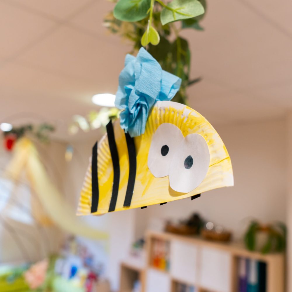 Nahaufnahme einer von Kindern gebastelten Biene aus einem gelb bemalten, gestreiften Pappteller mit großen, weißen Augen und einem blauen Krepppapier-Detail, die von der Decke hängt. Die Dekoration unterstreicht die naturpädagogische Ausrichtung und die Förderung der Kreativität im hellen Gruppenraum.